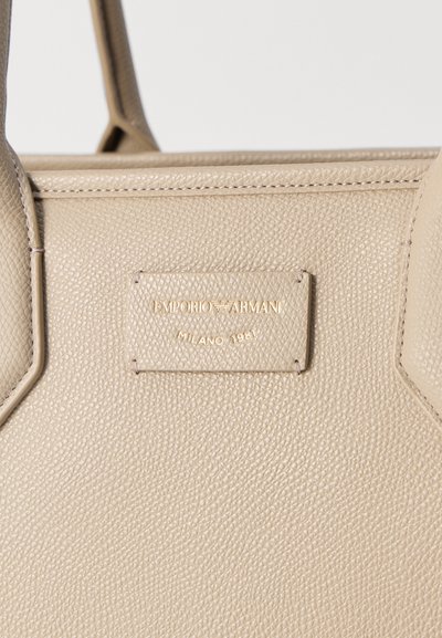 Μπεζ δερμάτινη τσάντα με ανάγλυφη ετικέτα που γράφει "EMPORIO ARMANI MILANO 1981" και δύο δερμάτινες λαβές.