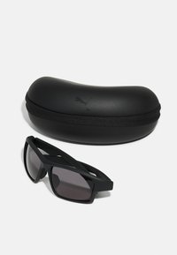 Schwarze Sonnenbrillen mit dunklen Gläsern und einem stabilen, eckigen Rahmen liegen neben einem eleganten, ovalen schwarzen Etui mit einem Logo.
