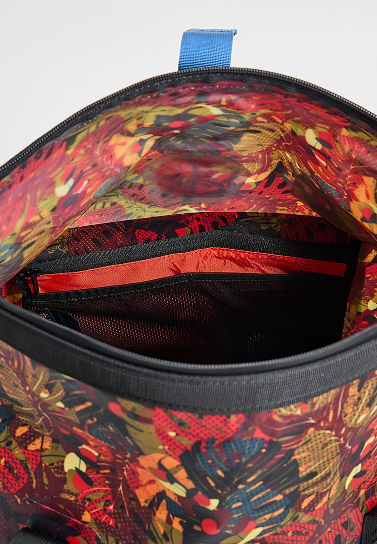 Zaino con motivo di foglie tropicali colorate, aperto per mostrare l'interno con tasca a rete con zip nera e rossa e maniglia blu.