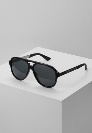 Sorte oversized solbriller med mørke linser, rund form, tykke glansfulde rammer og subtil branding på stængerne. Teksturede detaljer på armene.