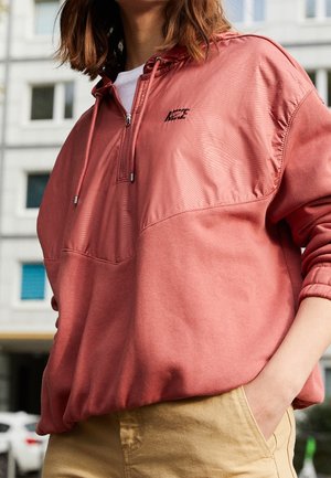 Persona con una sudadera con capucha de media cremallera en color rosa polvo con un pequeño logo en el pecho, pantalones beige, de pie al aire libre en un entorno urbano.