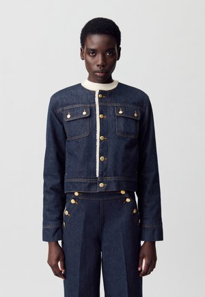 Veste en denim au design court, boutons dorés et col crème contrastant. Elle présente des poches poitrine doubles et une coupe structuré.