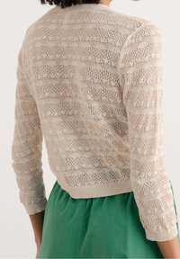 Cárdigan de punto color beige claro con patrón de calados, mangas 3/4 y dobladillo acanalado. Lleva puesto sobre una prenda verde sólida.