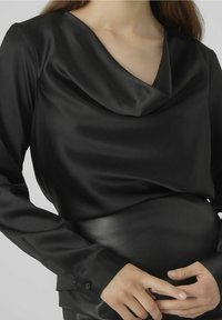 Femme portant un chemisier en satin noir avec un décolleté drapé et des manches longues, les mains doucement posées au niveau de la taille.