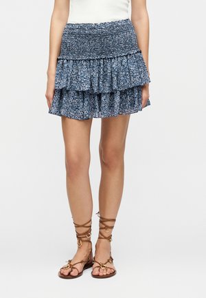 Kobieta ubrana w niebieską mini spódniczkę wielowarstwową w kwiaty ze ściągaczem w pasie oraz brązowe sandały na sznurowadła na jednolitym białym tle.