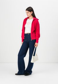Rode geribbelde zip-up hoodie, witte cropped T-shirt met zonnenafbeelding, hoge taille blauwe flared jeans, zwarte flats en kleine witte handtas.