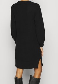 Robe en maille noire à manches longues, col rond, fentes latérales et tissu texturé. Le modèle arrive juste au-dessus des genoux.