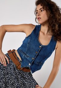 Denim vest in dark blue with scoop neckline, five buttons, and two front pockets. Paired with a patterned black and white skirt and a brown belt.