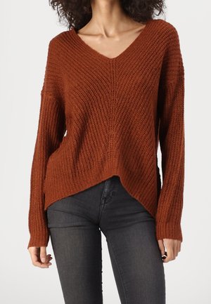 Mujer vistiendo un suéter de punto color óxido con cuello en V, combinado con jeans ajustados de color gris oscuro, sobre un fondo blanco liso.