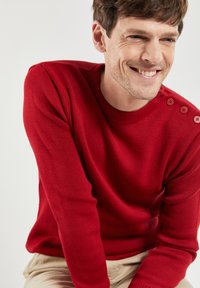 Roter gestrickter Pullover mit Rundhalsausschnitt und drei Knopfakzenten an der Schulter. Glatte Textur, figurschnitt Design. Heller Hintergrund.