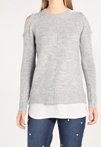 Grijze gebreide trui met uitsneden bij de schouders, geribbelde manchetten en zoom. Draag het over een witte gelaagde blouse en combineer het met blauwe spijkerbroek.