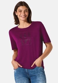 T-shirt à manches courtes violet en tissu doux, avec un design graphique en texte noir. Le col est rond et la coupe est décontractée.