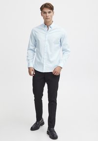 Helles blaues langärmeliges Hemd mit Knopfleiste und spitzem Kragen, kombiniert mit schwarzen schmal geschnittenen Hosen und schwarzen Slippern. Einfaches Design, glatte Textur.