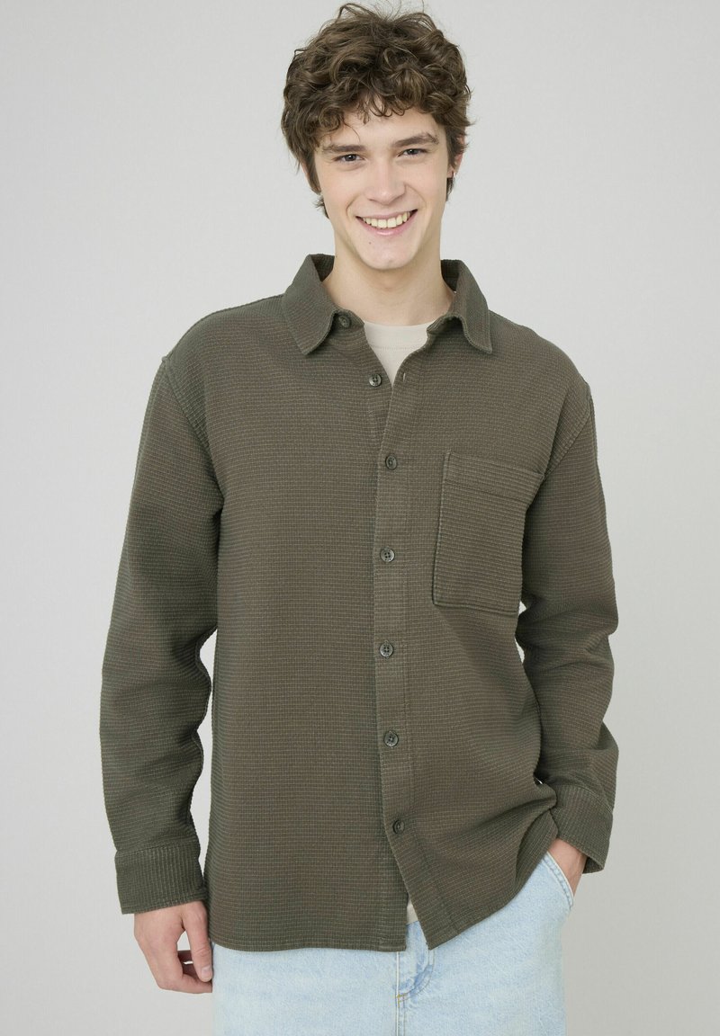 Chemise verte foncée à boutons, avec une finition texturée, à manches longues, poche poitrine unique et coupe décontractée, portée au-dessus d'un t-shirt clair.