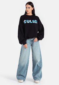 Czarna bluza z luźnym krojem, z turkusowym abstrakcyjnym napisem; zestawiona z jasnoniebieskimi dżinsami o szerokich nogawkach i kremowymi sneakersami.