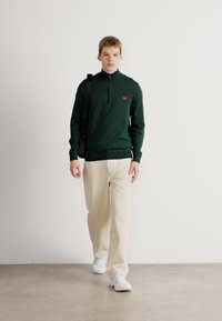 Dunkelgrüner Pullover mit Reißverschlusskragen, versehen mit einem kleinen roten Logo, kombiniert mit hellbeigen Hosen und weißen Sneakers. Glatte Stoffoberfläche.