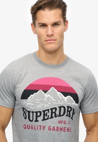 Gray T-shirt featuring a graphic design with mountains, a circular pink sunset, and the text "SUPERDRY MFG. CO QUALITY GARMENTS" in bold black.