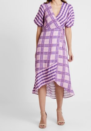 Kvinde iført en lilla og pink plaid slå-om kjole med puffede ærmer og asymmetrisk kant, kombineret med beige ankelrem-sandaler.
