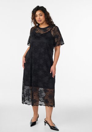 Žena v čiernych midi šatách s kvetinovou čipkou a krátkymi rukávmi, nosí ich cez čierne spodné šaty, doplnené čiernymi lodičkami s otvorenou pätou a špičkou na bielom pozadí.