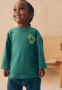 Grøn langærmet t-shirt med en gul and-grafik på brystet, kombineret med blå jeans med striber, stående foran en træbaggrund.