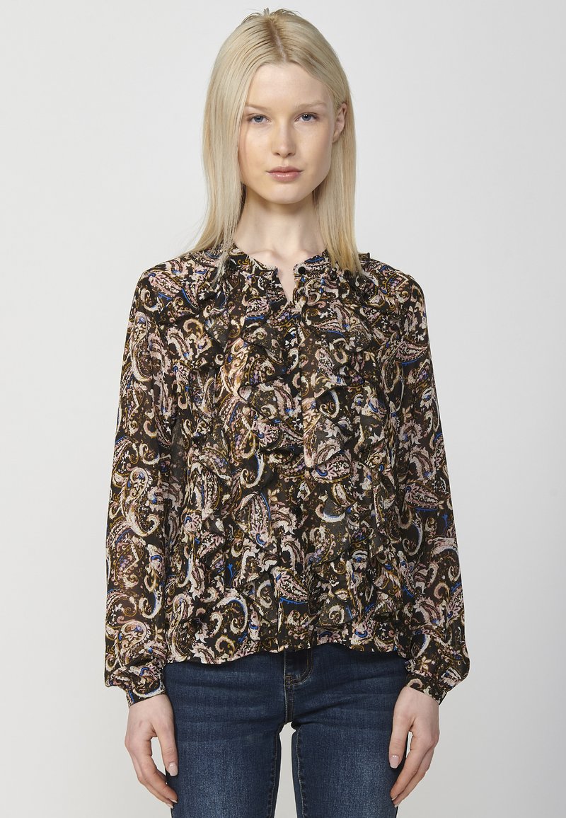 Bluză neagră cu model, având un design paisley multicolor și detalii cu volane pe decolteu, asortată cu blugi strâmti albaștri.