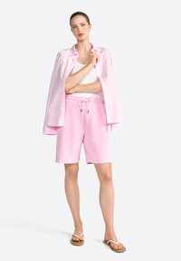 Chemise surdimensionnée rose clair, débardeur blanc et short rose assorti avec cordon de serrage. Le mannequin porte des tongs beiges ; tissu doux et coupe décontractée.