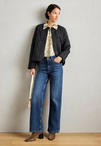 Giacca nera imbottita sopra un top chiaro a fantasia, abbinata a jeans blu a vita alta e a gamba larga e stivaletti marroni con tacco. È in mano una borsa beige.