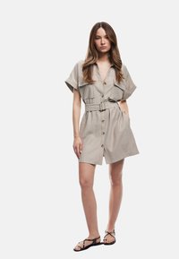 Abito beige con cintura, maniche corte, chiusura a bottoni frontale, due tasche sul petto e una vestibilità comoda, abbinato a sandali neri flat.