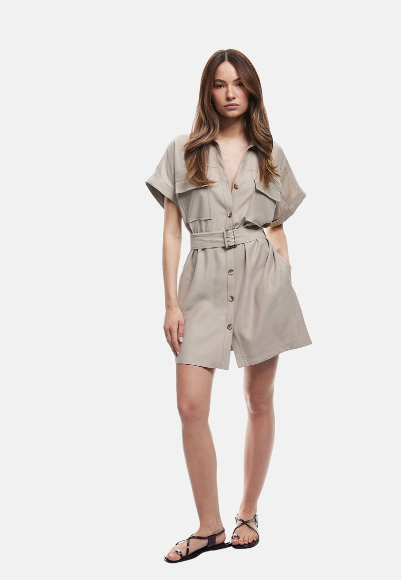 Abito beige con cintura, maniche corte, chiusura a bottoni frontale, due tasche sul petto e una vestibilità comoda, abbinato a sandali neri flat.