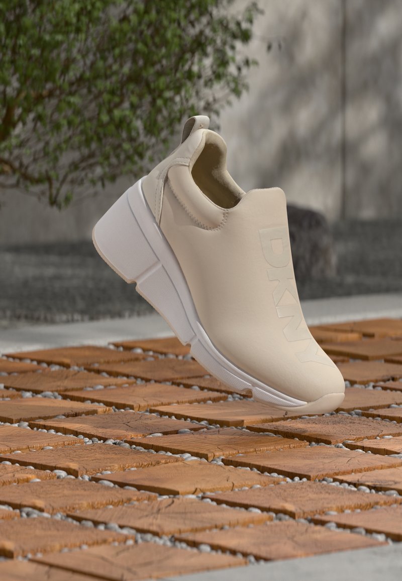 Μπεζ αθλητικό παπούτσι τύπου slip-on με χοντρή λευκή σόλα και ανάγλυφο «DKNY» στη πλευρά, τοποθετημένο πάνω σε ξύλινα πλακάκια με κενά γεμάτα χαλίκι σε εξωτερικό χώρο.