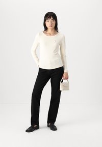 Cremfarbener, gerippter Langarmoberteil mit Rundhalsausschnitt, kombiniert mit schwarzen, geraden Hosen und einer kleinen weißen Handtasche. Schwarze Slipper.