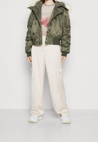 Chaqueta bomber corta en color verde oliva con capucha forrada de pelo, combinada con pantalones de pierna ancha en color blanco roto y zapatillas blancas. Presenta múltiples bolsillos.