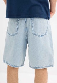 Shorts de mezclilla azul claro con un corte recto, dos bolsillos traseros y un dobladillo ligeramente deshilachado, combinados con una camisa de un tono oscuro de marino.