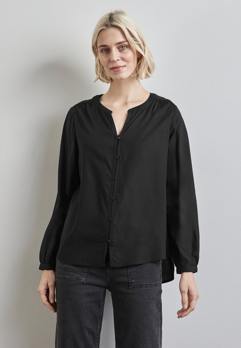 Blusa nera a maniche lunghe con scollatura a V, chiusura frontale con bottoni e spalle arricciate. Tessuto liscio con una leggera lucentezza.