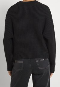 Person som bär en svart grovstickad tröja och mörkgrå jeans med synliga bakfickor, som står mot en enkel ljus bakgrund.