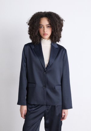 Navy satin blazer with notched lapels, two front pockets, and a single button closure, paired with a cream, ruffled turtleneck underneath.