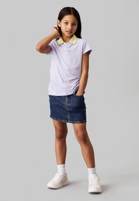 Polo de color púrpura claro con cuello amarillo, falda mini de mezclilla y zapatillas blancas. El diseño incluye un logo en el pecho y un detalle en el bolsillo.