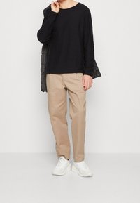 Svart långärmad skjorta, avslappnad passform, tillsammans med beige byxor och vita sneakers. En grå vadderad jacka över axeln.