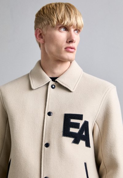 Μπεζ μάλλινη ζακέτα με γιακά, μαύρα κουμπιά και μαύρο φelt λογότυπο "EA" στο αριστερό στήθος. Λείος υφή, χαλαρός σχεδιασμός.