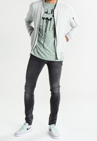 Svetle šedá bomber bunda přes mátově zelené tričko s černým grafickým potiskem, kombinováno s tmavými slim kalhotami a mátovými teniskami Nike.