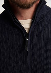 Marineblauwe gebreide zip-up trui met ribbeltextuur en opstaande kraag; voorzien van een zwarte rits en een subtiele accent bij de hals.