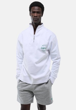 Homme portant un sweat-shirt blanc à fermeture éclair quart de zip avec un texte vert et un short gris clair, debout les mains dans les poches sur un fond uni.