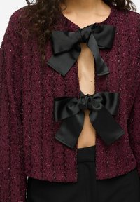 Burgundy kort kardigan med texturerad tyg, med två stora svarta satinsnören vid halsringningen och en öppen frontdesign.