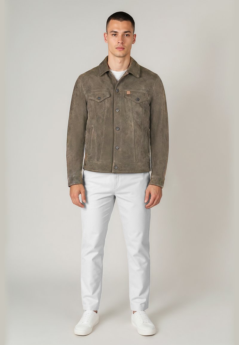 Mann steht und trägt eine braune Wildlederknopflederjacke, ein weißes Hemd, weiße Hose und weiße Sneakers vor einem einfarbigen hellen Hintergrund.