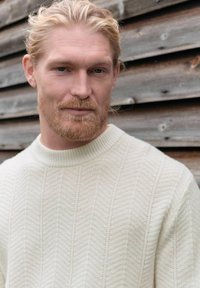 Pull crème tricoté avec un motif chevrons texturé et un col rond côtelé. Porté par un mannequin devant un fond en bois.