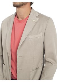 Beige kavaj i lätt material, enfärgad med slagkragar, två fickor och en dekorativ detalj på försidan. Bärs över en korallfärgad t-shirt.