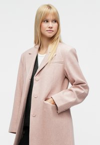 Casaco de lã cor de rosa claro com design ajustado, apresentando três botões, bolsos laterais e colarinho com recorte. O tecido texturizado confere uma aparência suave.