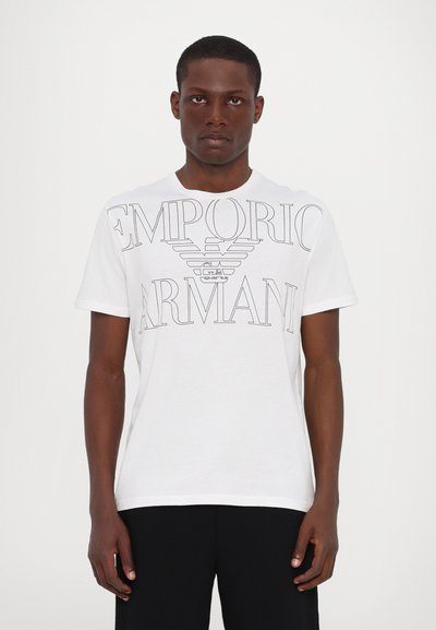 Camiseta de algodón blanca con un gran logo impreso "EMPORIO ARMANI" junto con un emblema de águila. Mangas cortas, cuello redondo, ajuste relajado.