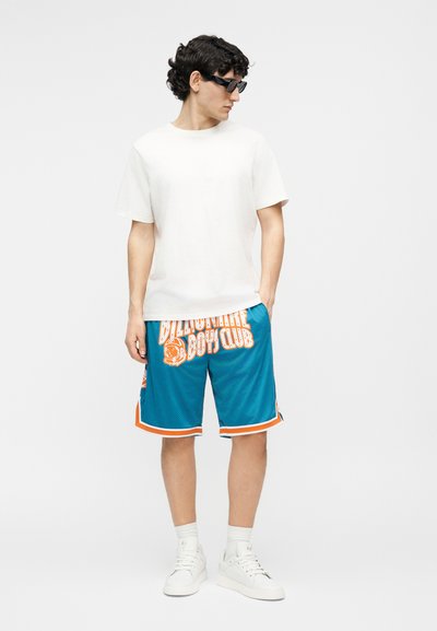 Ember fekete napszemüveget visel, egyszerű fehér pólót, kék és narancssárga "Billionaire Boys Club" sortot, fehér zoknit és fehér tornacipőt, áll.