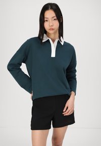 Tyrkysová polokošeľa s dlhým rukávom a bielym golierom, s tromi gombíkmi, skombinovaná s čiernymi šortkami. Materiál sa zdá byť mäkký a má voľný strih.